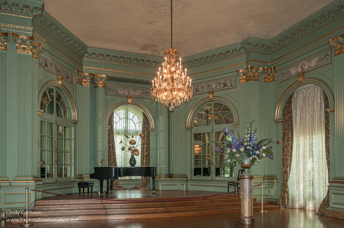 Filoli&rsquo;s ballroom stage area before a 2014 renovation, with large windows, a grand piano, dramatic flower arrangements, and an elaborate chandelier. 