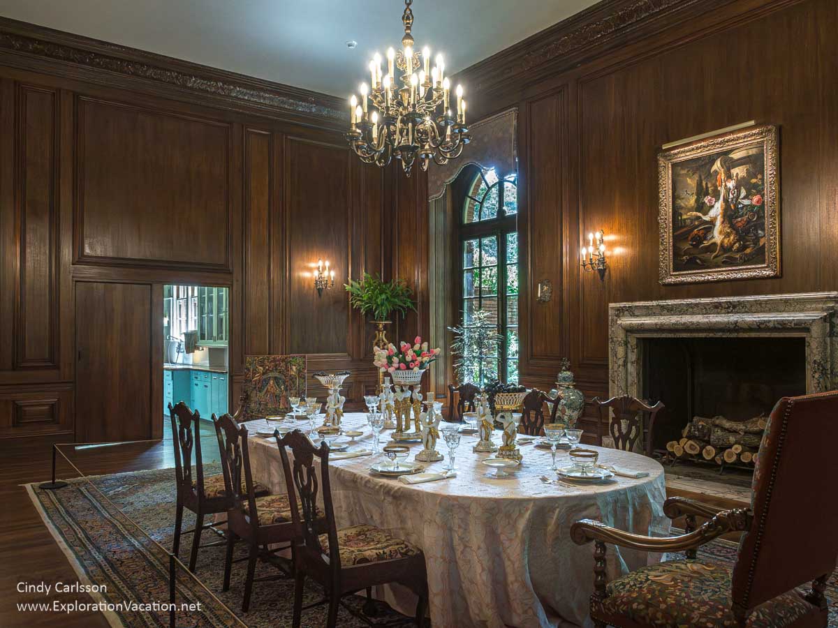 The dining room in Filoli with the dining room table set with china and crystal, a fireplace, paintings on the walls, and the butler&rsquo;s pantry visible through an open door.