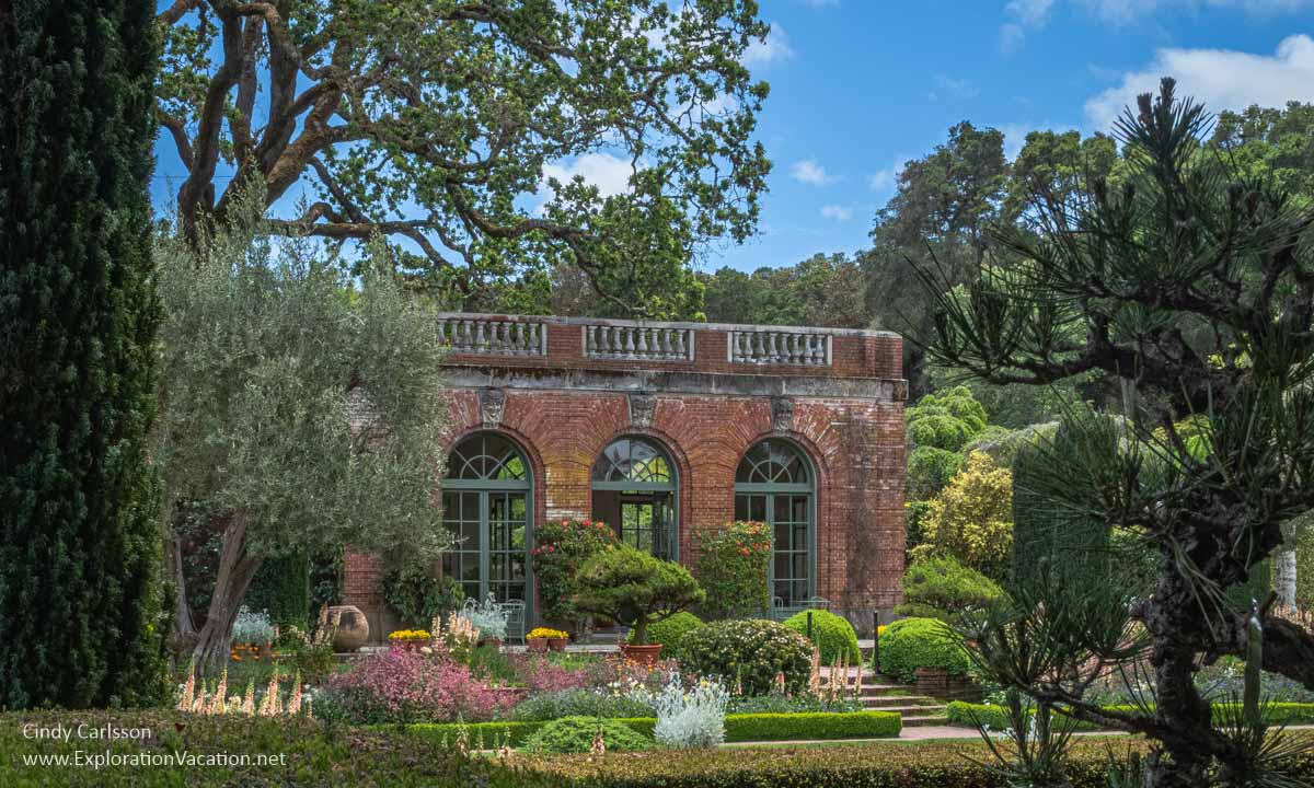 A small brick garden house with large windows and surrounded by plants.  