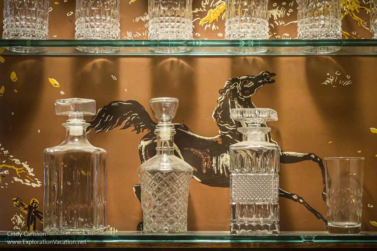 Glass decanters and glasses on glass shelves with a prancing horse on the wallpaper behind them. 