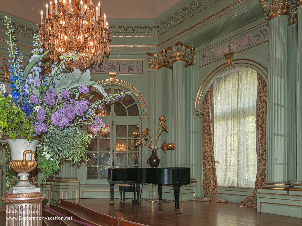 A flower arrangement under a chandelier and a grand piano by a window in an elegant ballroom. 