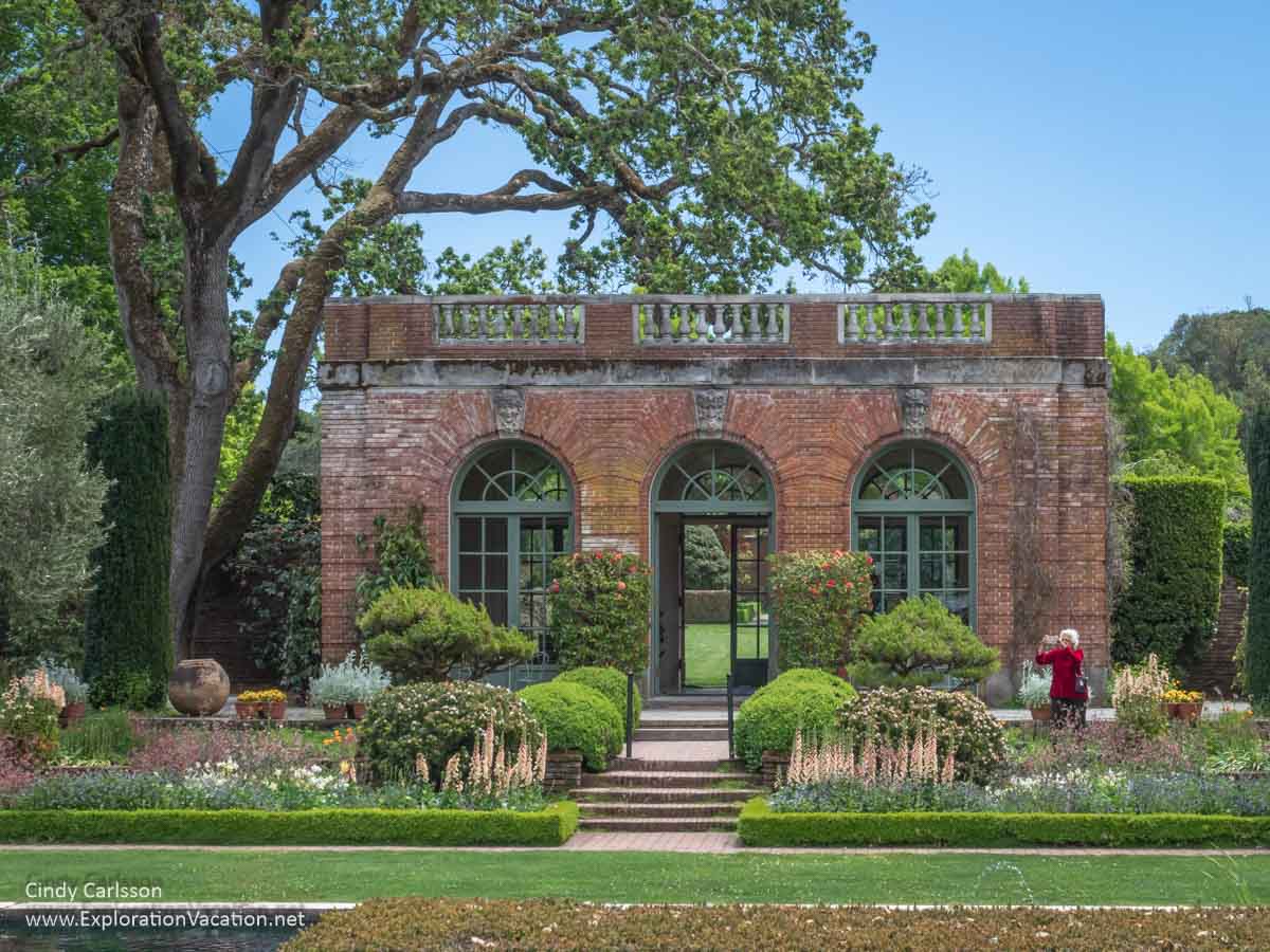 A small brick garden house with large windows and surrounded by gardens.