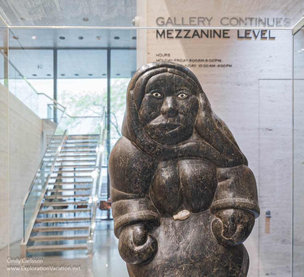 Photo of an Inuit sculpture by Johnny Inukpuk of a woman, with stairs in the background and a sign for the Mezzanine at the TD Gallery of Indigenous Art.