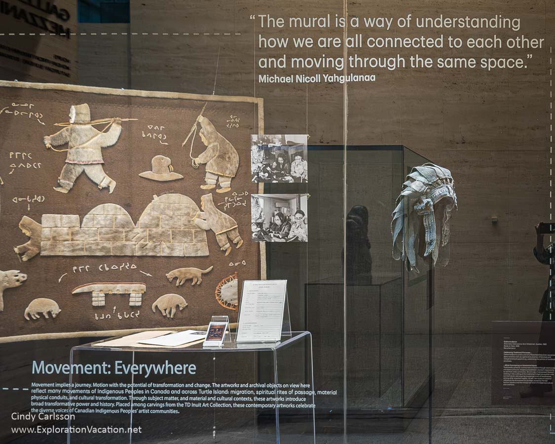 Photo of a display case with a traditional-style headdress made from old jeans by Atikamekw Nehirowisiw artist Catherine Boivin and a vintage textile by Inuit arts Mary Cookie and Alice Margeyok displayed at the TD Gallery of Indigenous Art.