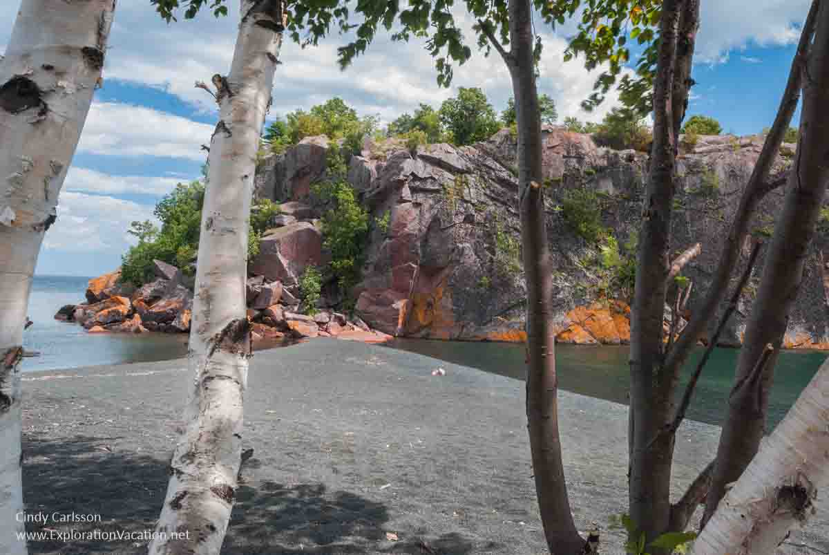 Photo of a black sand beach and rock formations seen through a few birch trees along Lake Superior in Silver Bay on Minnesota’s North Shore © Cindy Carlsson at ExplorationVacation.net
