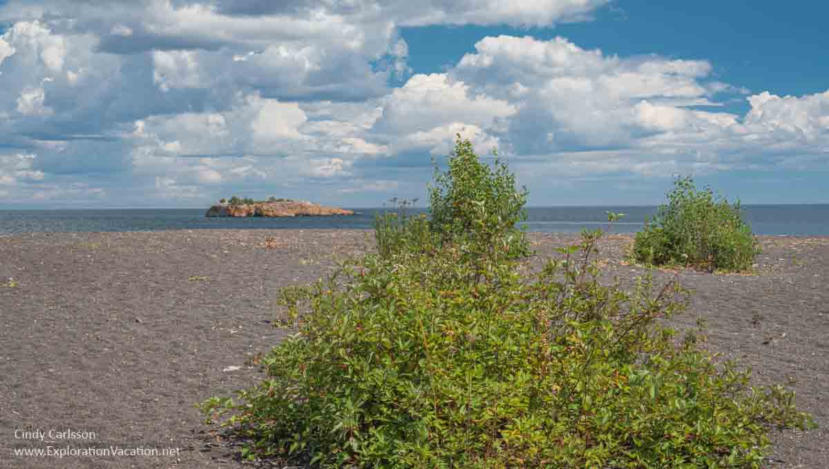 Photo of a black sand beach with a few small shrubs and an off-shore island on Lake Superior in Silver Bay Minnesota © Cindy Carlsson at ExplorationVacation.net