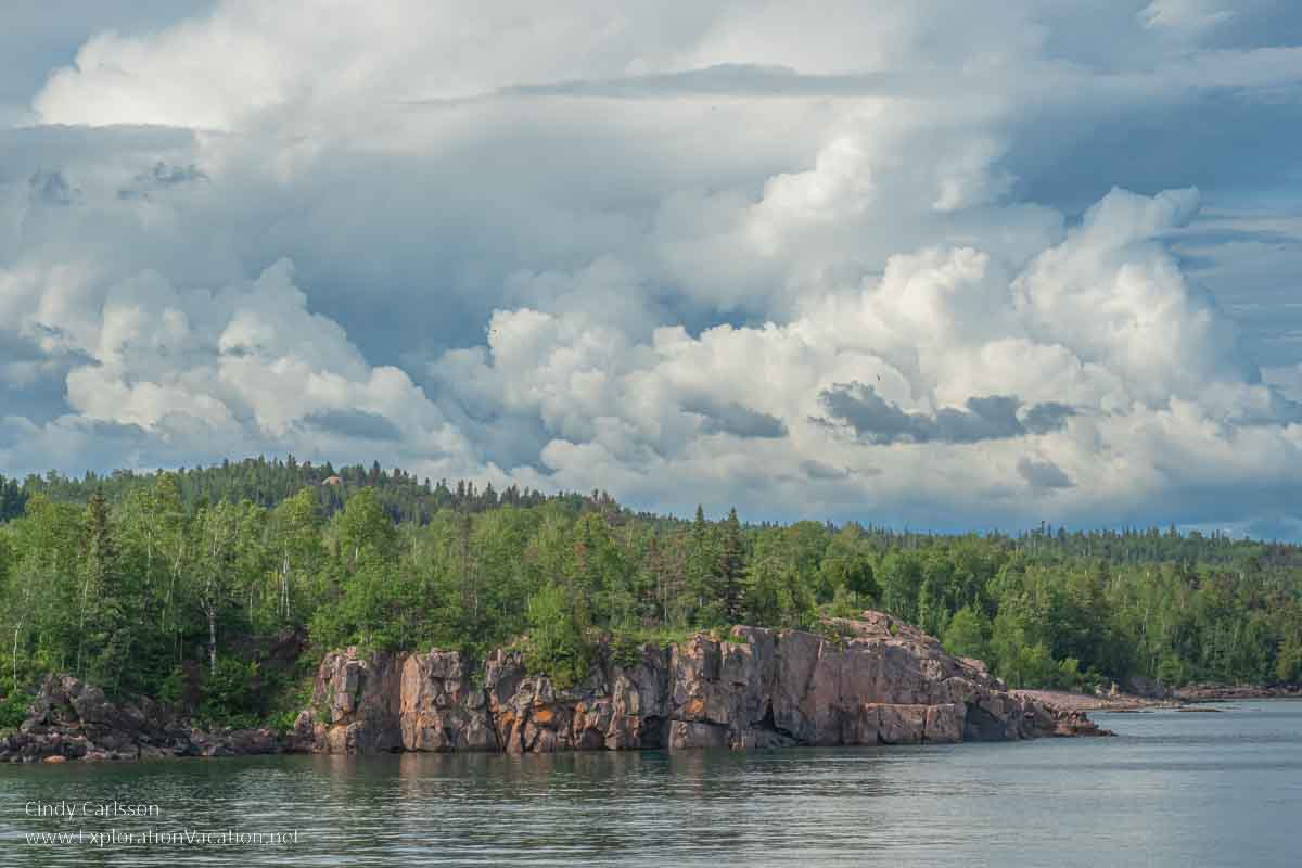 Photo of a rock formation with forested hills behind it and dramatic clouds in the sky at Black Beach on Lake Superior in Silver Bay Minnesota © Cindy Carlsson at ExplorationVacation.net