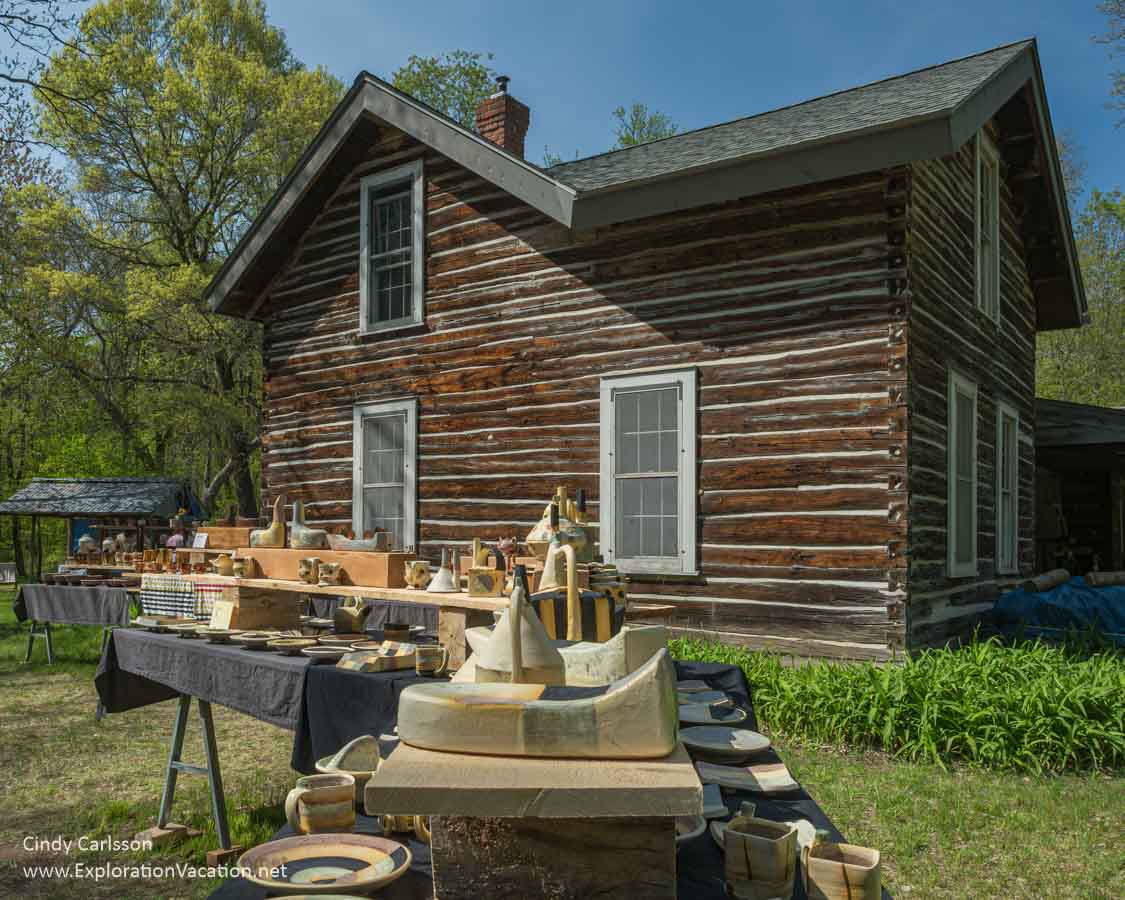 Photo of a log house with pottery by Mike Helke set out on tables in the yard during the 2024 St. Croix Valley Pottery tour in Minnesota. (Photo &copy; Cindy Carlsson on ExplorationVacation.net)