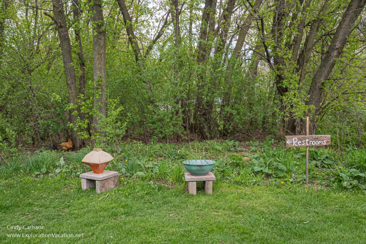 A restroom sign, pottery, and a chicken on the grass by a hedgerow. 