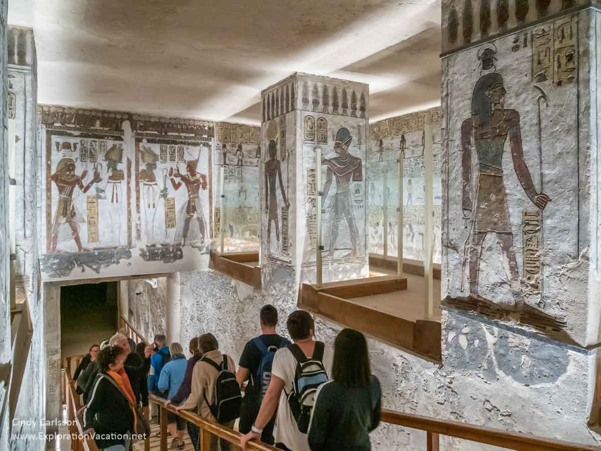 People walking down a ramp below large paintings of pharaohs and gods inside a tomb