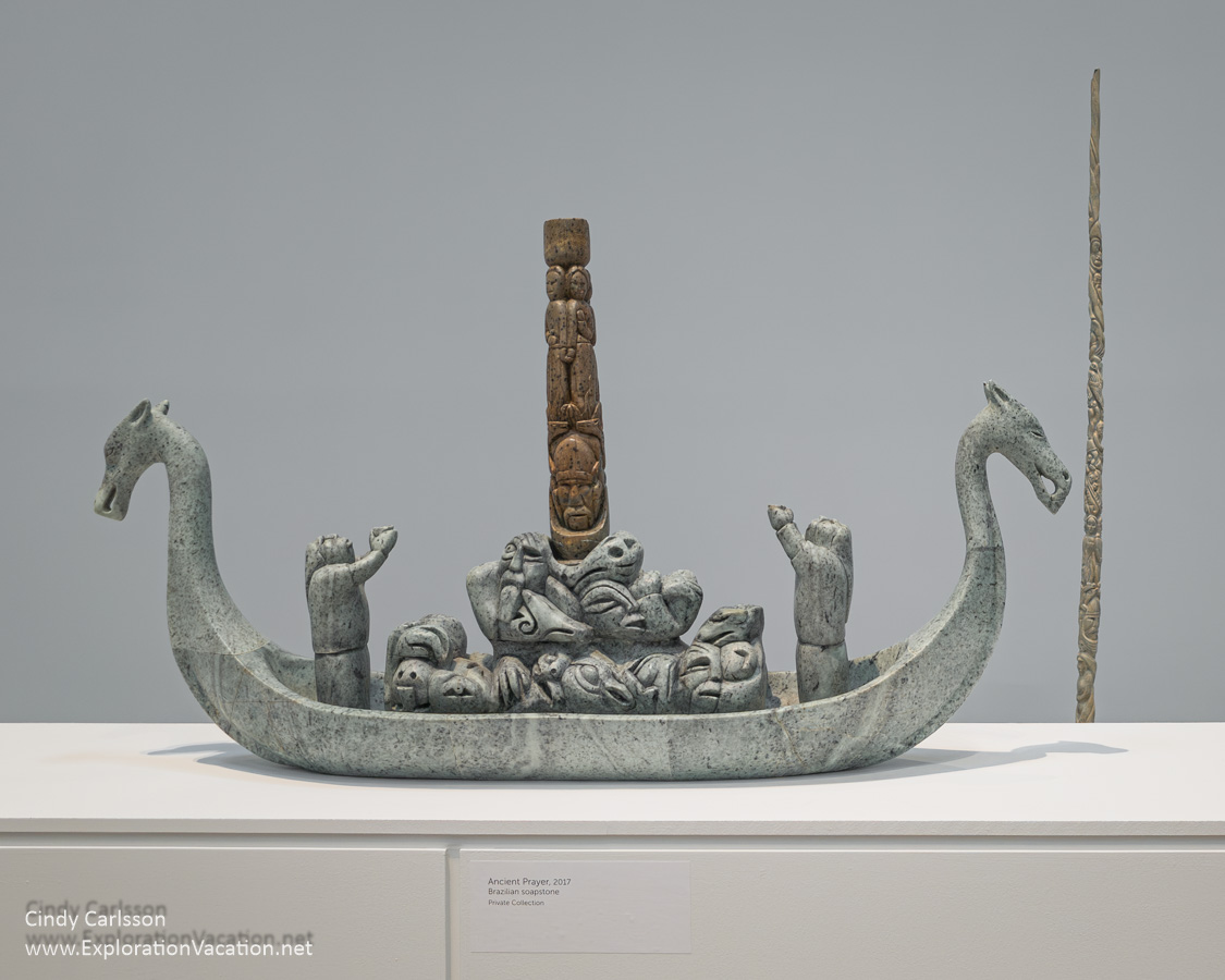 A stone carving of a Viking ship with figures making offerings to a pillar in mid ship and a bronze sculpture that looks like a carved narwhal tusk both by Abraham Anghik Ruben.