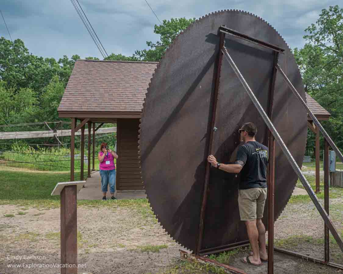 Photo of a huge round saw blade with a man standing behind it and a woman taking his picture in Quarry Park and Nature Preserve near St Cloud Minnesota. Text and photos © Cindy Carlsson ExplorationVacation.net