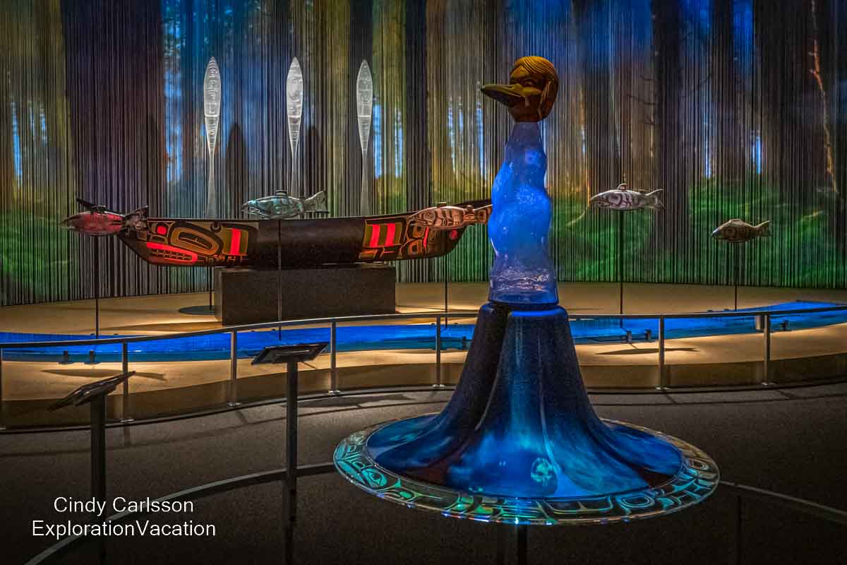 photo of a raven transforming into a human in front of a Tlingit canoe with raised paddles and swimming salmon (all created in glass and colorfully lit) by Tlingit artist Preston Singletary for "Raven and the Box of Daylight" at the National Museum of the American Indian Washington DC © Cindy Carlsson at ExplorationVacation.net