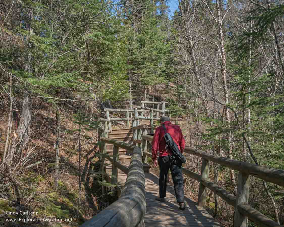 Photo of a man standing on a wooden boardwalk in the woods with railings and stairs visible in the distance at the Caribou Falls State Wayside on Minnesota&rsquo;s North Shore &copy; Cindy Carlsson at ExplorationVacation.net