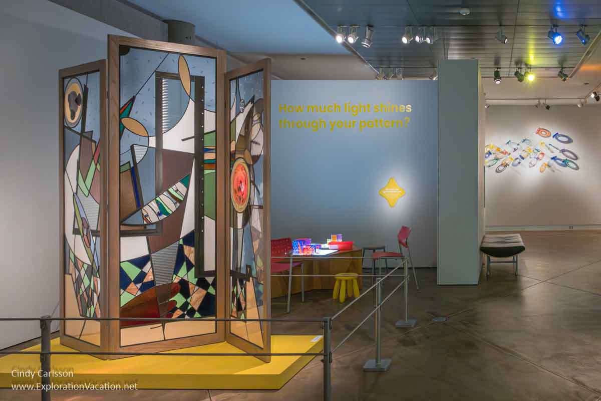 Photo of a gallery with a large glass folding screen, a table with items intended for children to play with, and another glass art piece visible in the background in the exhibit &ldquo;Illuminate: Glass Art for Early Learners&rdquo; at the Museum of Glass in Tacoma, Washington in July 2023 &copy; Cindy Carlsson at ExplorationVacation.net