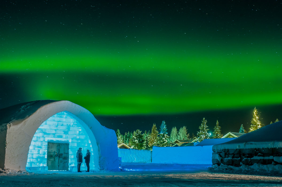 Photo of people standing by the entrance to Icehotel 27 at night with the northern lights overhead. Photo &copy; Asaf Kliger for the ICEHOTEL www.icehotel.com 