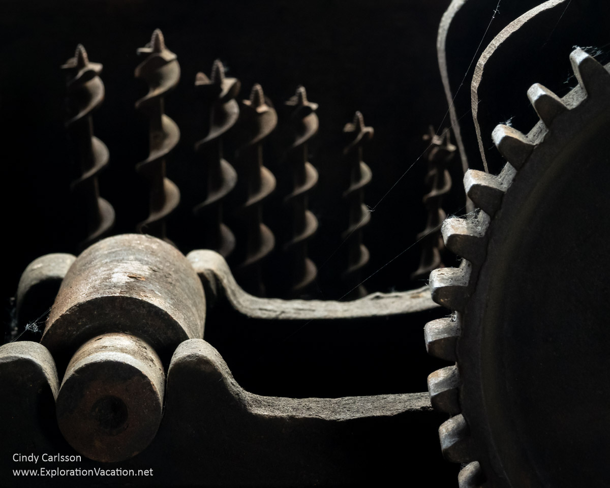 An almost abstract photo of shadowy equipment with a large gear and an out-of-focus set of very large drill bits inside the Bally Blacksmith Shop in Grand Marais, Minnesota &copy; Cindy Carlsson at ExplorationVacation.net