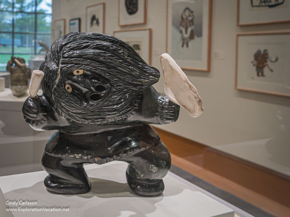 A drum dancer by Uriash Puqiqnaq with prints on the wall behind.