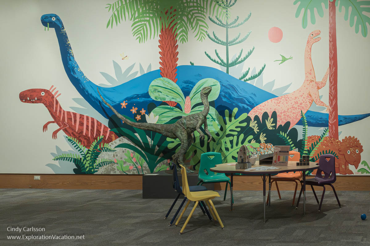 Children at a table in front of a wall with a dinosaur mural at the Dennos.