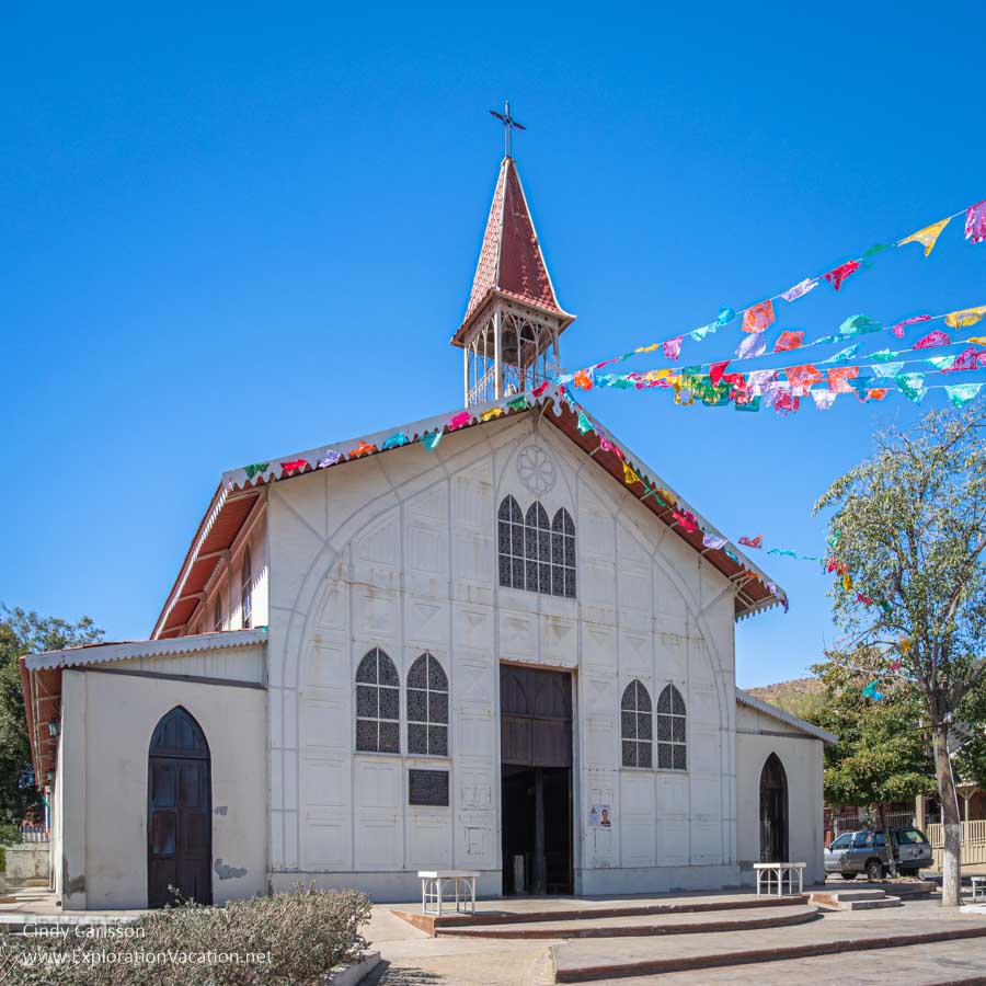 A simple white metal church with a red roof and steeple with strips of colorful flags suspended from the steeple. 