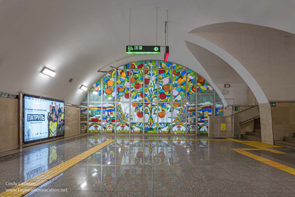 An Almaty subway station hall with a colorful glass mural of an apple tree at one end.