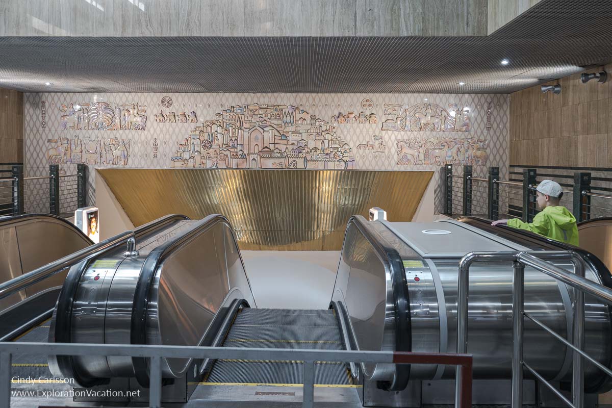A large ceramic mural with what appears to be a Central Asian city and caravans above an escalator.