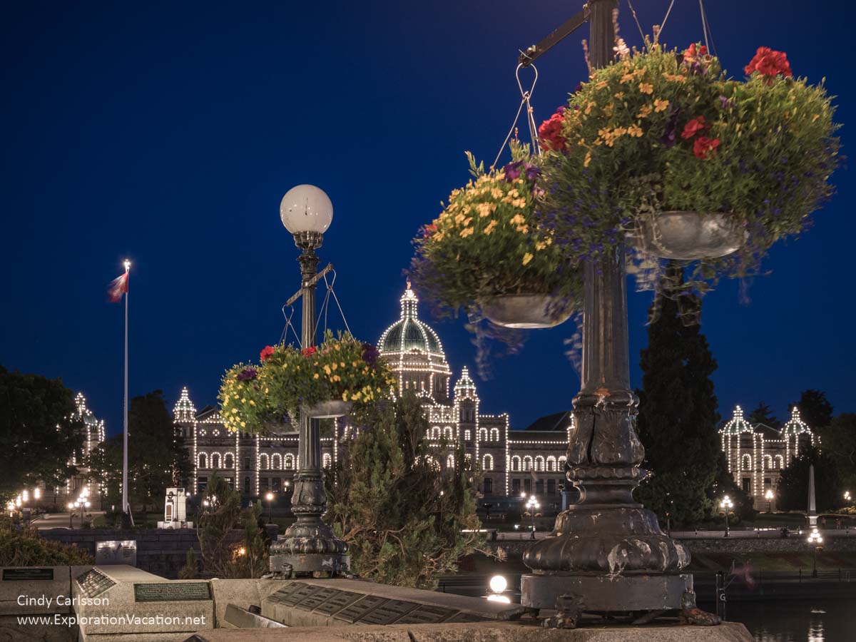 Night photo of the walkways near the parliament in Victoria, British Columbia, with flower baskets and the parliament building outlined with lights in the background.