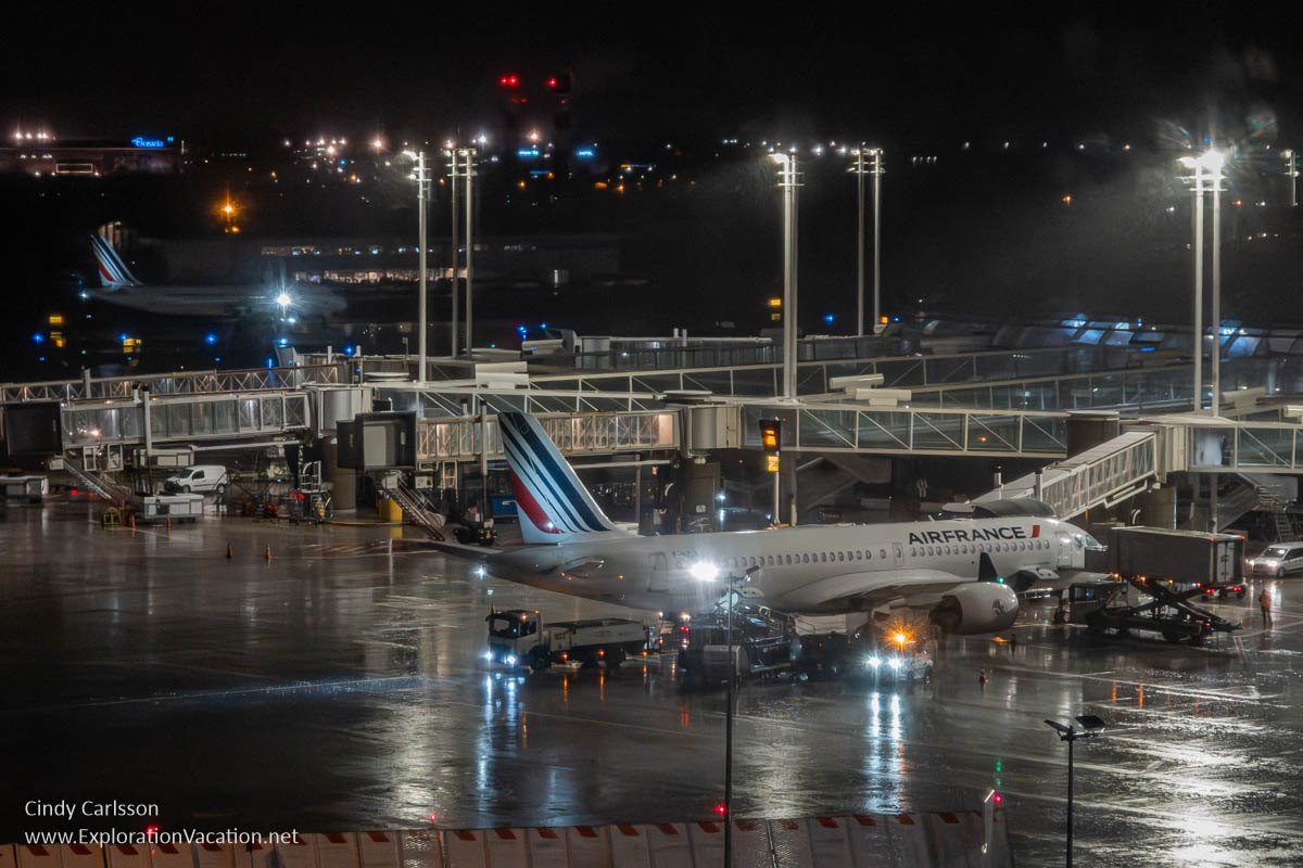 An Air France plane and support vehicles at a gate with another in the background on a rainy night at Charles de Gaulle airport.