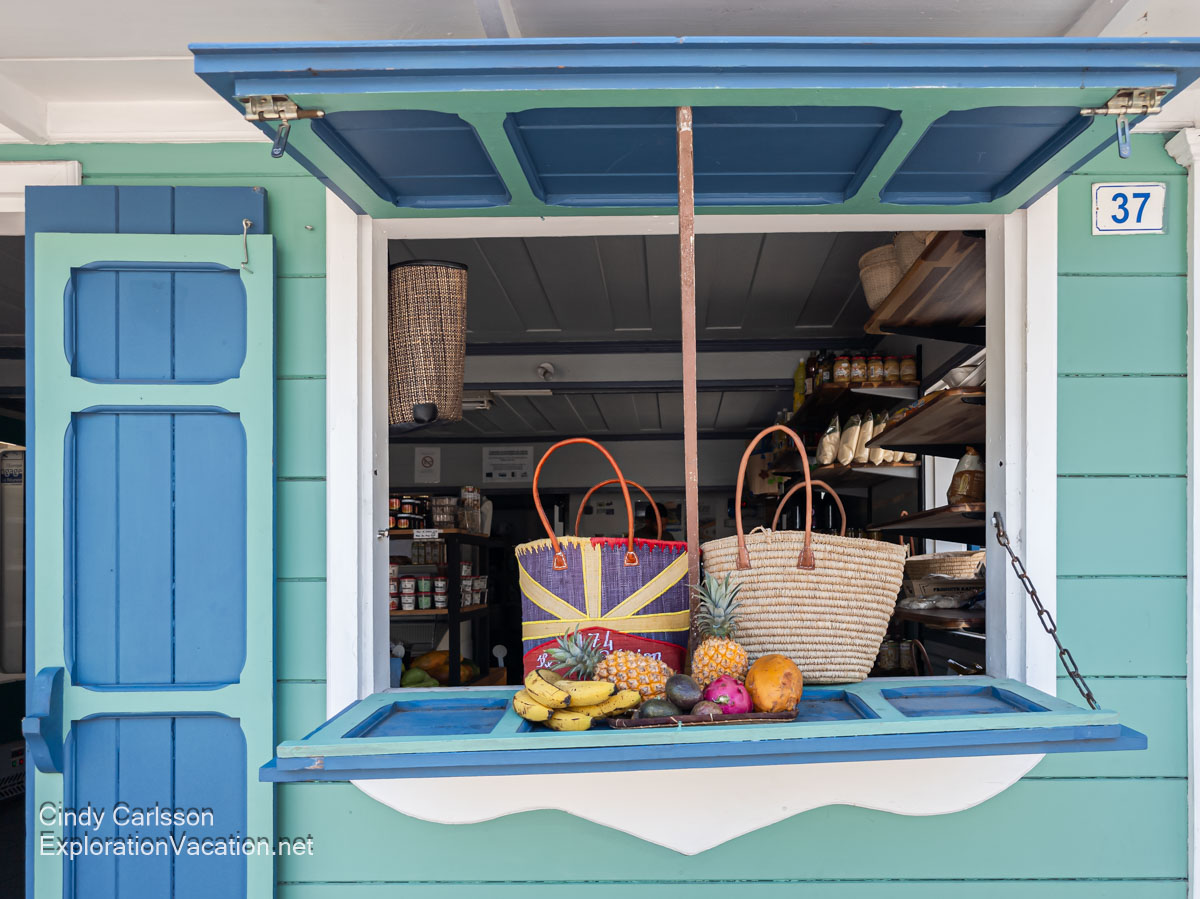 A grocery store window display with woven straw market baskets and neat piles of bananas, pineapple, mango, and other fruits.