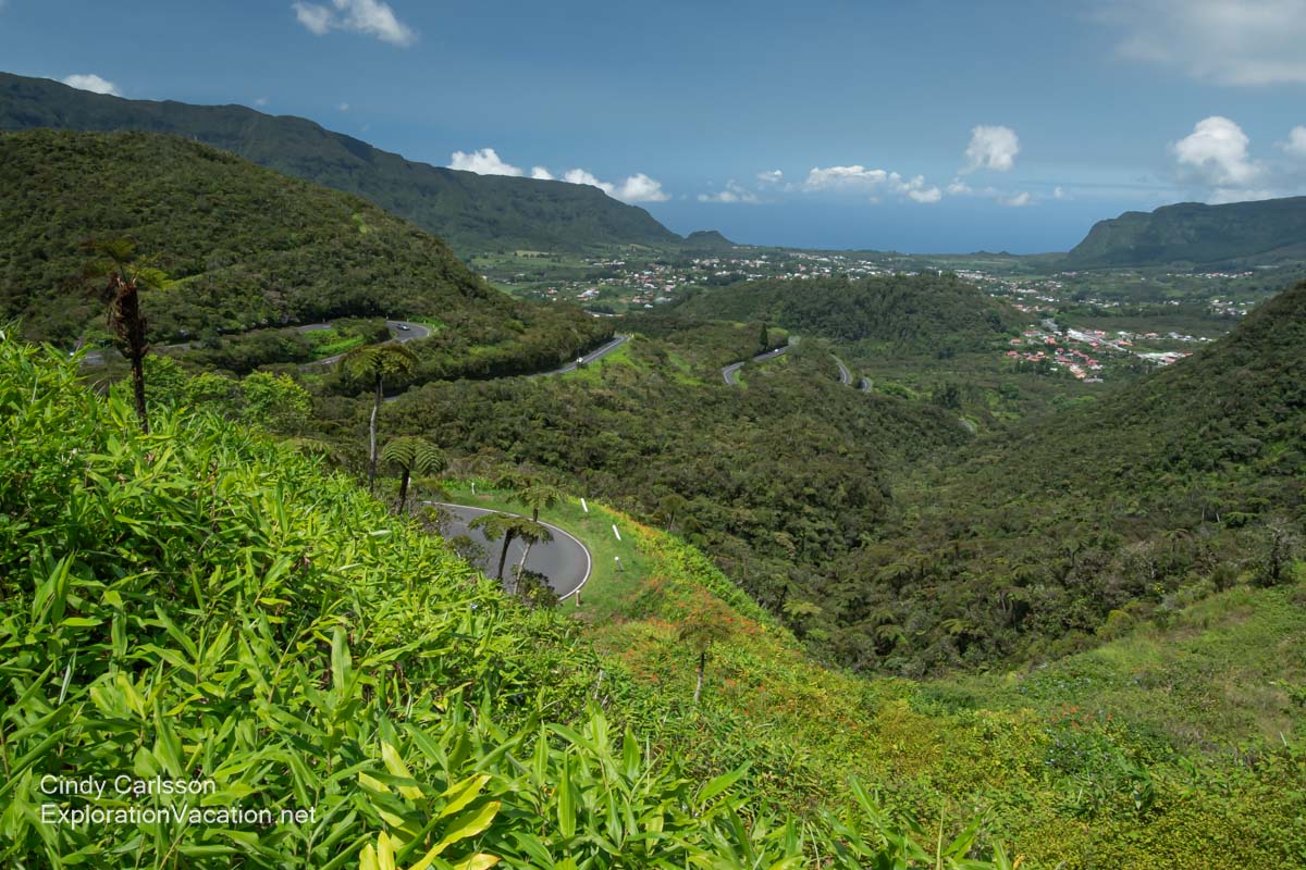 Overview of an expansive forested valley with many switchbacks on the road through it and urban areas visible in the distance.