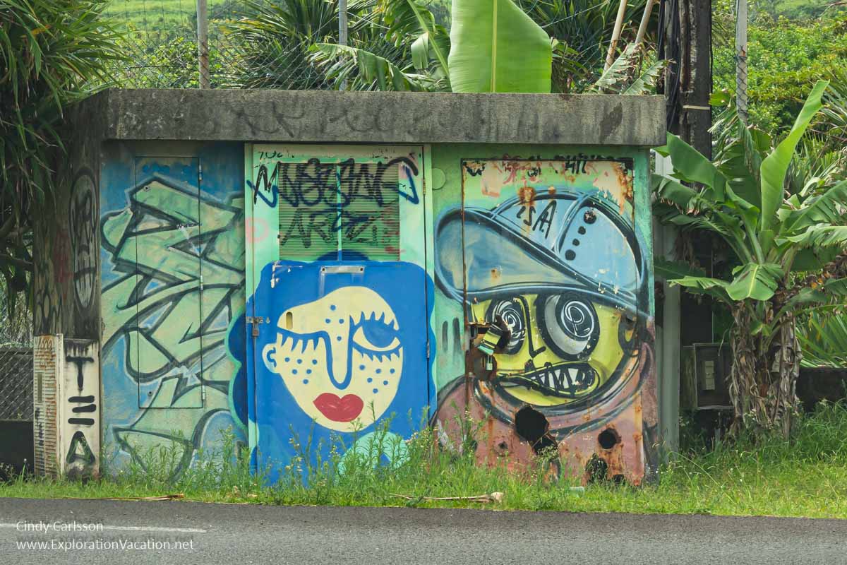 A shed with two faces and graffiti painted on it along a roadside. 