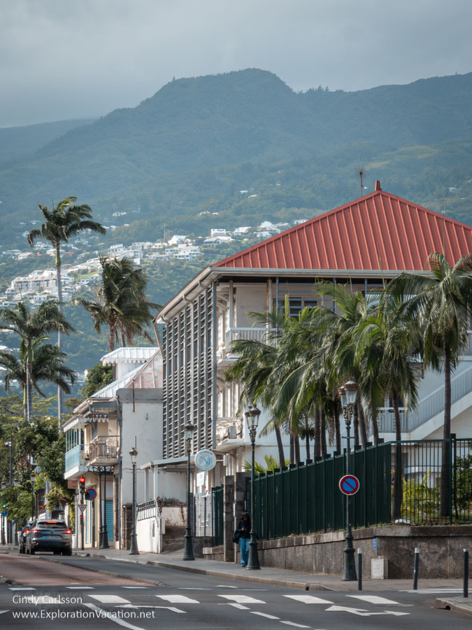 Street scene with balcony-lined buildings, palm trees, and mountains rising in the background.