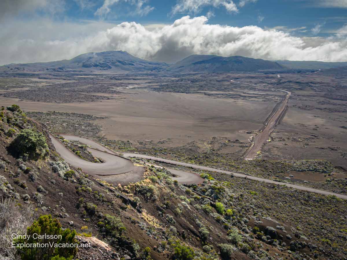 Looking down to a twisting road, a barren plain, and volcanic mountains beyond.