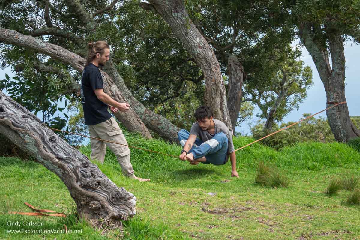 Two young men, one sitting on a slack line suspended between trees with the sea visible far in the distance.