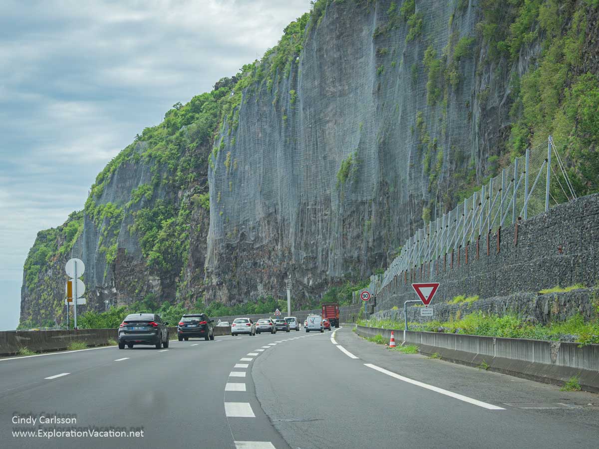 Multi-lane highway at the foot of a mountainside draped in netting to stop rockfalls.