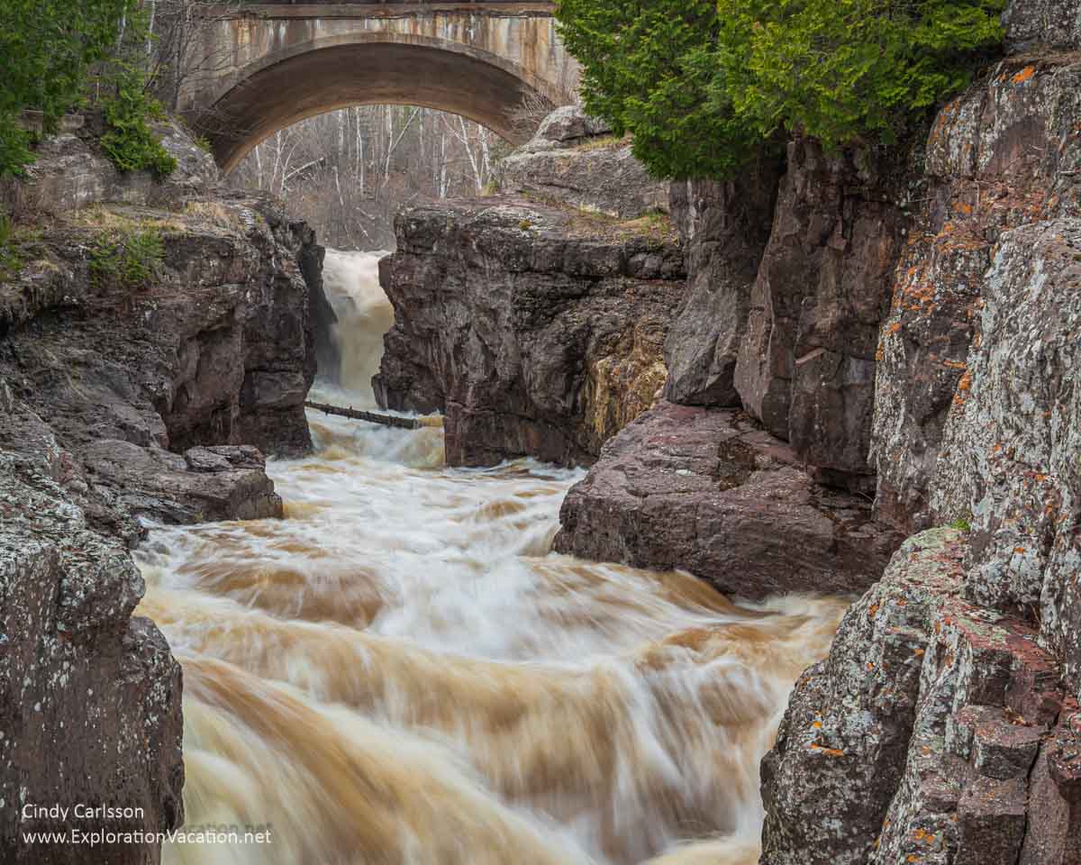 A roaring river tumbling through an arch and between high rock cliffs.