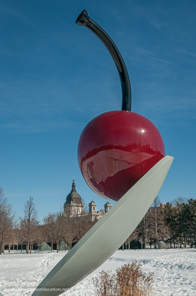 A very large sculpture of a cherry perched on a spoon in winter, with the Basilica in the background.