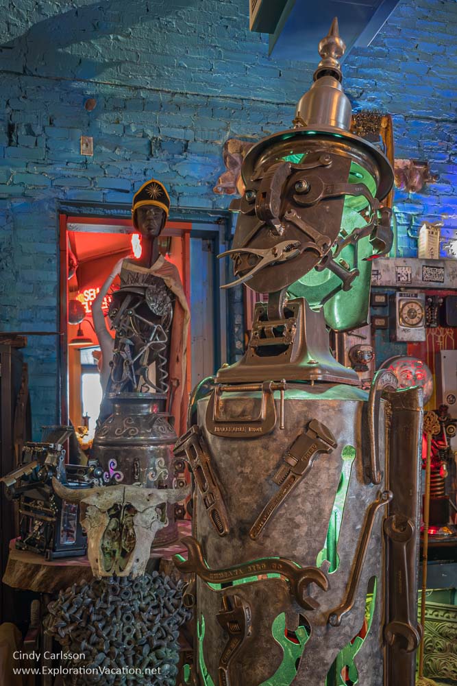 A humanoid figure made from old tools and scrap metal with glowing green light emanating from it while an oddly arrayed caped superhero of some sort stands in the background.