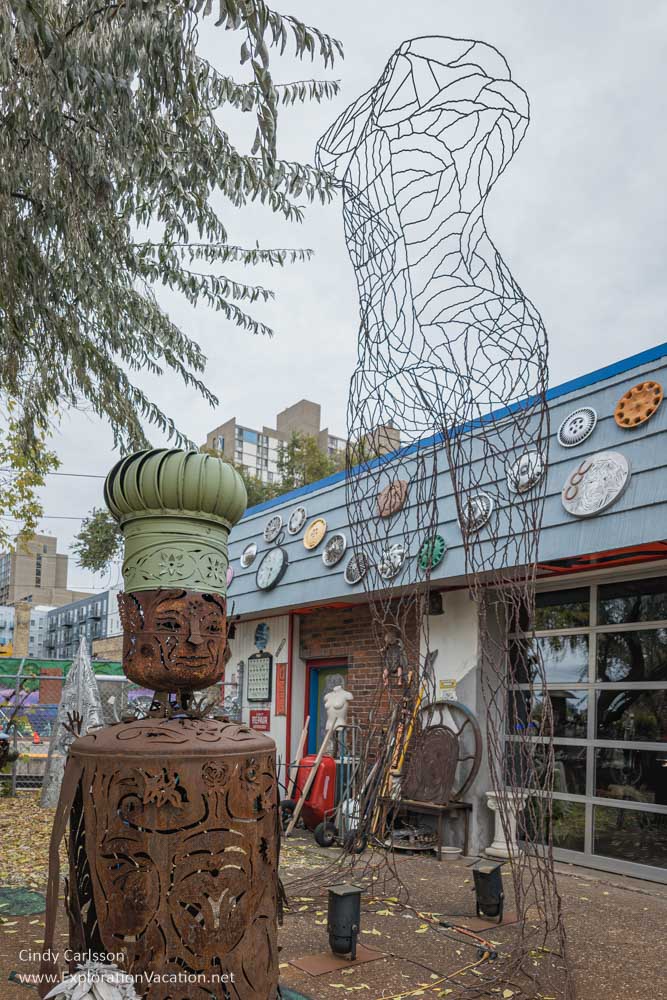 A metal sculpture wearing an air vent as a chef's hat, a towering wire outline of a woman, and other art and found objects with the building's glass garage door in the background.